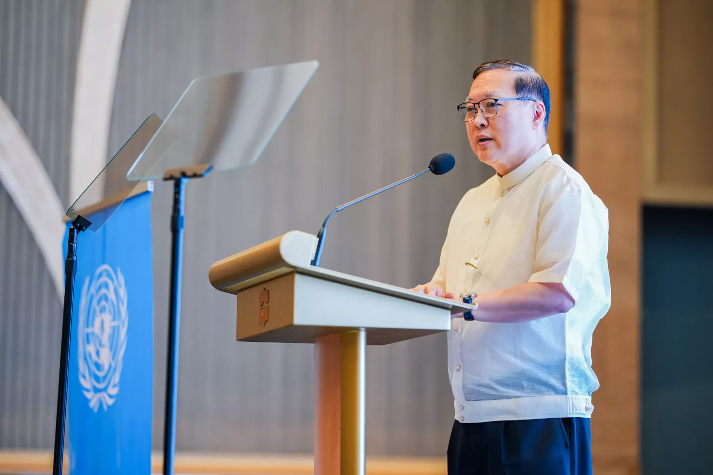 Asiatic man standing on a podium and delivering a message during an event, with a United Nations flag beside him