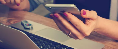 A woman looking at her smartphone while sat at her desk in front of her laptop