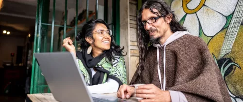 A smiling woman and a man sit at an outdoor café, chatting over a laptop. A colorful mural with a flower and guitar is behind them.