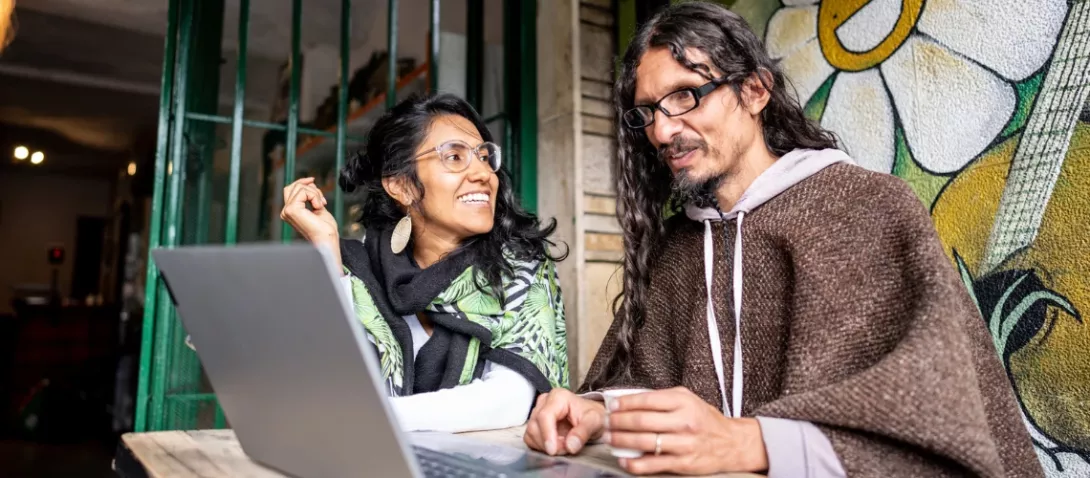 A smiling woman and a man sit at an outdoor café, chatting over a laptop. A colorful mural with a flower and guitar is behind them.